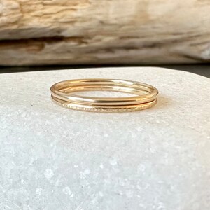 May include: Three gold stacking rings. The bottom ring has a hammered texture. The rings are on a white surface.