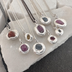 Peut inclure: Une collection de colliers avec pendentifs en pierres précieuses. Chaque collier présente une pierre brute de forme irrégulière sertie dans un pendentif en métal argenté. Les pierres précieuses sont de couleurs variées, dont le rouge, le rose, le jaune et le bleu, et sont suspendues à de fines chaînes.