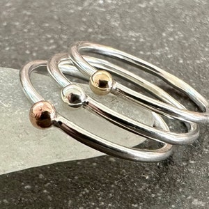 May include: Three silver rings with a small ball at the end of each ring. The balls are different colours: gold, silver, and rose gold.