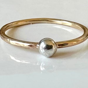May include: A delicate ring featuring a gold band with a hammered texture and a small, silver-coloured sphere at the centre. The ring's design is simple and elegant, suitable for everyday wear. The ring is a piece of jewellery.