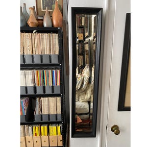 May include: A tall, rectangular mirror with a black frame is mounted on a white door. The mirror reflects a room with various objects, including a shelf filled with books and decorative vases. The mirror's frame is approximately 60 inches tall.
