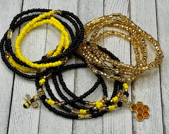 May include: A set of black, yellow, and gold beaded waist beads with bee and honeycomb charms. The beads are arranged in a layered pattern, with the black beads forming the base and the yellow and gold beads layered on top.