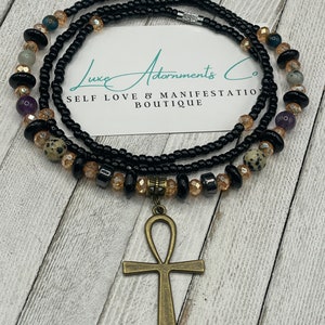 May include: A black beaded waist bead with a gold ankh pendant. The beads are interspersed with small clear and brown beads. The waist bead is on a wooden surface.