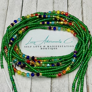 May include: A collection of green waist beads with colorful accents. The beads are a deep green, with strands of gold, red, yellow, blue, and purple beads interspersed throughout. The image includes a white card with the text "Luxe Adornments Co. SELF LOVE & MANIFESTATION BOUTIQUE."