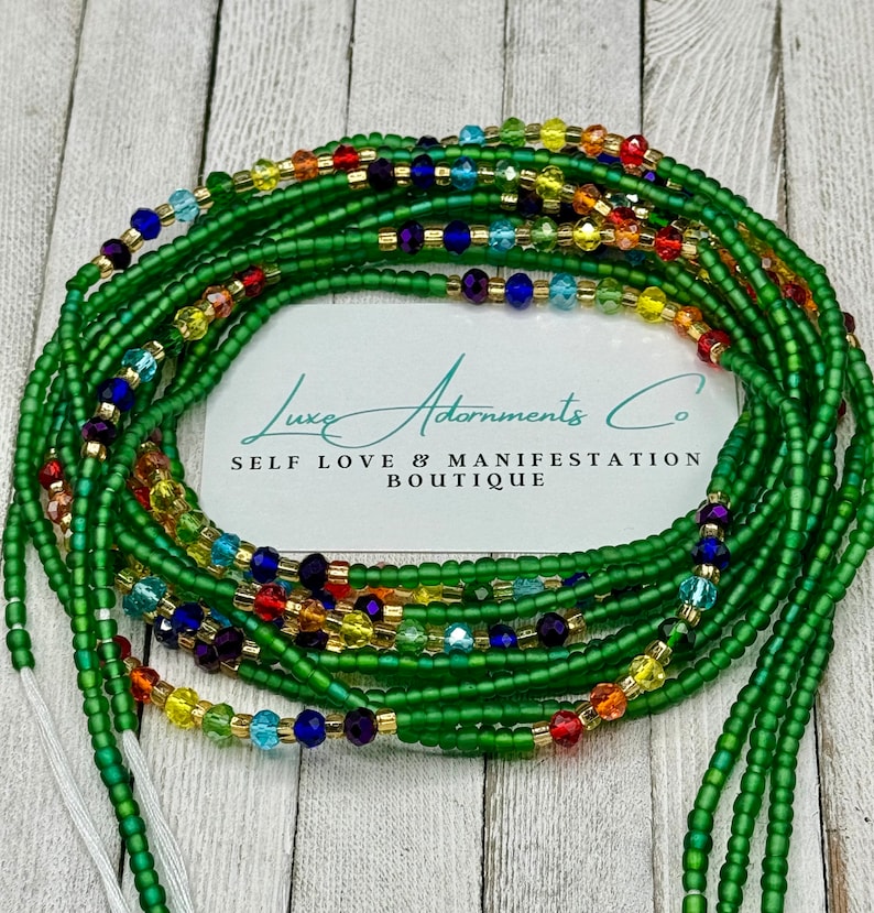May include: A collection of green waist beads with colorful accents. The beads are a deep green, with strands of gold, red, yellow, blue, and purple beads interspersed throughout. The image includes a white card with the text "Luxe Adornments Co. SELF LOVE & MANIFESTATION BOUTIQUE."