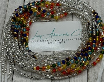 May include: A collection of beaded waist beads with a rainbow of colors, including red, orange, yellow, green, blue, and purple. The beads are strung on a silver-colored string. The image includes a white card with the text "Luxe Adornments Co. SELF LOVE & MANIFESTATION BOUTIQUE."