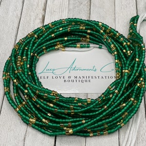 May include: A collection of green and gold waist beads. The beads are a deep green colour with gold accents. The beads are strung together on a white string. The image includes the text "Luxe Adornments Co. SELF LOVE & MANIFESTATION BOUTIQUE."