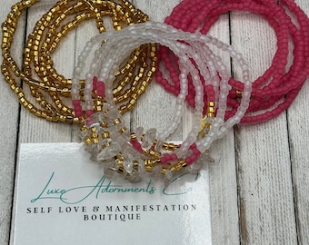 May include: A set of three waist beads, one gold, one white, and one pink. The beads are arranged in a circle and are on a wooden surface. The beads are made of small, round beads. The text "Luxe Adornments Co. Self Love & Manifestation Boutique" is on a white card below the beads.