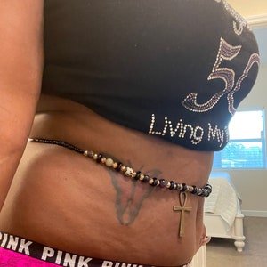 May include: A black waist beads with gold and purple beads, a gold ankh charm, and a butterfly tattoo on the wearer's abdomen. The beads are worn over a black shirt with rhinestones that reads "Living My Best Life" and a pink underwear with the word "PINK" repeated.