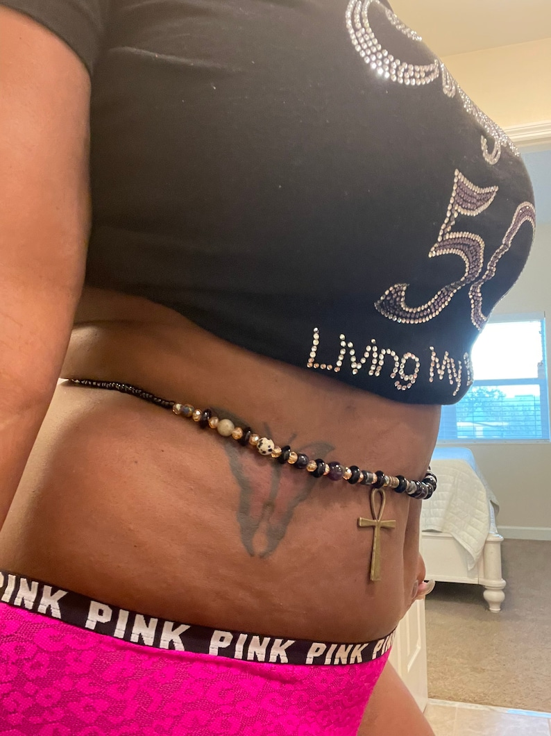 May include: A black waist beads with gold and purple beads, a gold ankh charm, and a butterfly tattoo on the wearer's abdomen. The beads are worn over a black shirt with rhinestones that reads "Living My Best Life" and a pink underwear with the word "PINK" repeated.