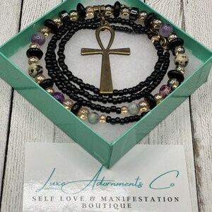 May include: A black box with a gold and black beaded necklace with an ankh pendant. The box has the text "Luxe Adornments Co. Self Love & Manifestation Boutique" on it.