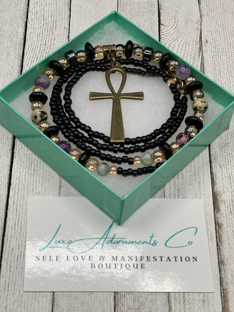 May include: A black box with a gold and black beaded necklace with an ankh pendant. The box has the text "Luxe Adornments Co. Self Love & Manifestation Boutique" on it.