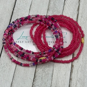 Puede incluir: Un conjunto de cuentas de cintura de color rosa y morado con detalles dorados. Las cuentas están dispuestas en círculo y apiladas unas sobre otras. Las cuentas están hechas de vidrio y tienen un acabado mate.