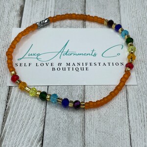 K&ouml;nnte beinhalten: Ein Regenbogen-Ankle-Armband mit goldener Akzent und einem silbernen Verschluss. Das Armband besteht aus orangefarbenen, roten, gelben, gr&uuml;nen, blauen, indigofarbenen und violetten Perlen. Das Armband befindet sich auf einem wei&szlig;en Hintergrund mit dem Text "Luxe Adornments Co. Self Love & Manifestation Boutique".