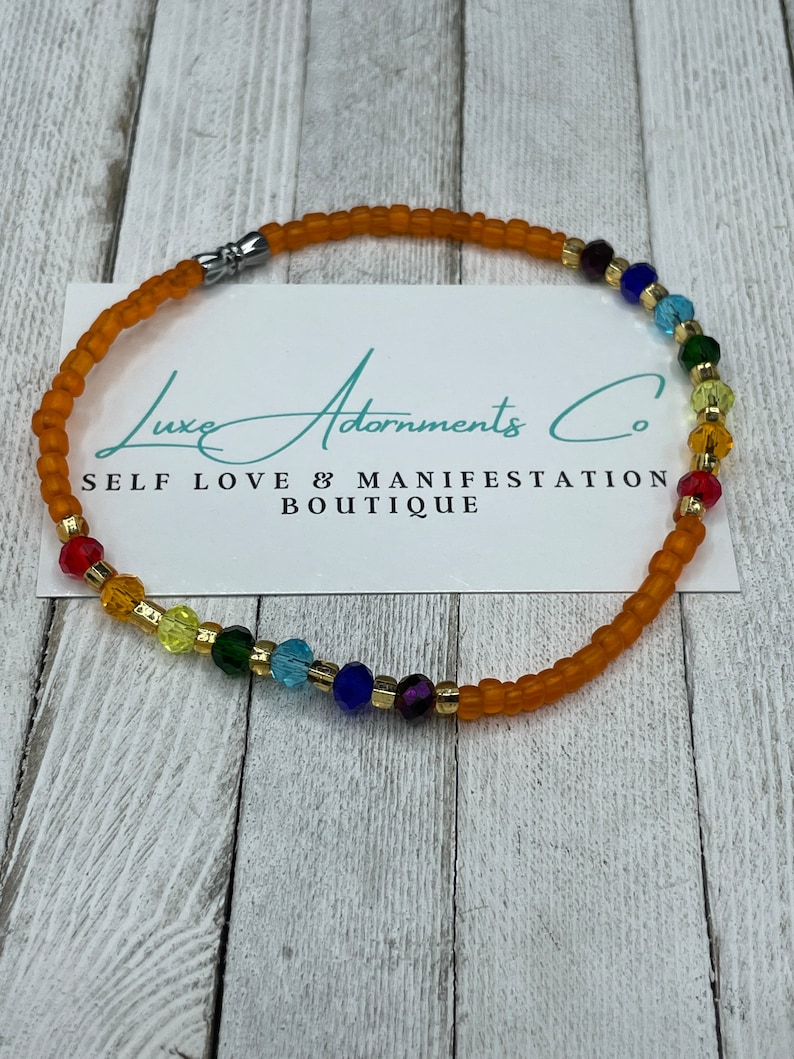 K&ouml;nnte beinhalten: Ein Regenbogen-Ankle-Armband mit goldener Akzent und einem silbernen Verschluss. Das Armband besteht aus orangefarbenen, roten, gelben, gr&uuml;nen, blauen, indigofarbenen und violetten Perlen. Das Armband befindet sich auf einem wei&szlig;en Hintergrund mit dem Text "Luxe Adornments Co. Self Love & Manifestation Boutique".