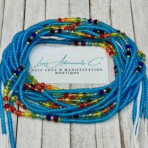 May include: A collection of light blue waist beads with colorful accents. The beads are arranged in a circular pattern, with a white card in the center that reads "Luxe Adornments Co. SELF LOVE & MANIFESTATION BOUTIQUE."