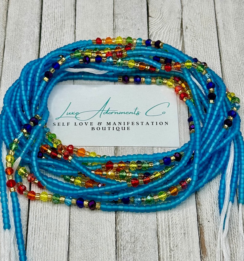 May include: A collection of light blue waist beads with colorful accents. The beads are arranged in a circular pattern, with a white card in the center that reads "Luxe Adornments Co. SELF LOVE & MANIFESTATION BOUTIQUE."