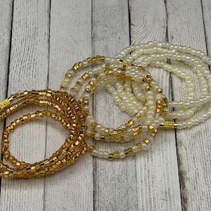 May include: Three strands of waist beads in gold and ivory colors. The beads are small and strung together, creating a delicate and elegant look. The gold beads have a metallic sheen, while the ivory beads have a pearlescent finish. The beads are arranged on a light wood grain surface.
