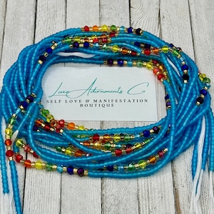 May include: A blue waist bead necklace with colorful glass beads. The beads are arranged in a rainbow pattern. The necklace is on a wooden surface with a white card that says "Luxe Adornments Co. Self Love & Manifestation Boutique".