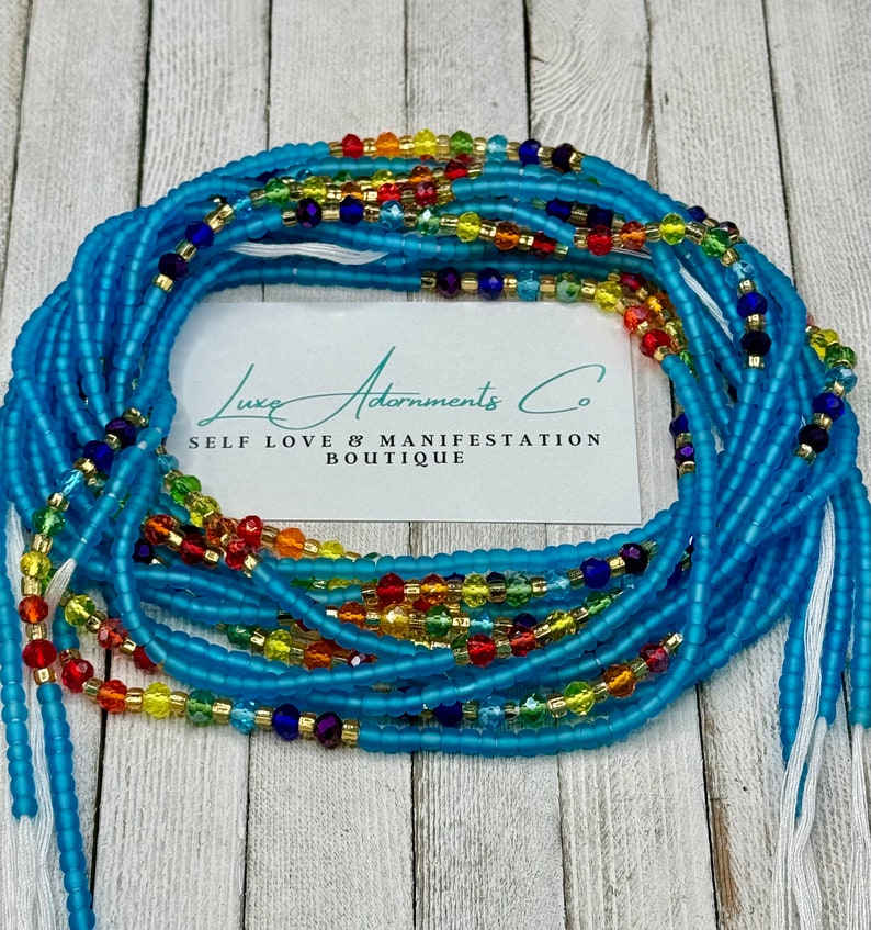 May include: A blue waist bead necklace with colorful glass beads. The beads are arranged in a rainbow pattern. The necklace is on a wooden surface with a white card that says "Luxe Adornments Co. Self Love & Manifestation Boutique".
