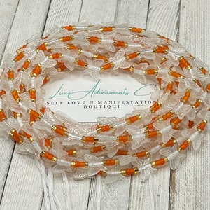 May include: A waist bead necklace made with clear butterfly beads and orange and gold beads. The beads are strung on a white cord. The necklace is coiled and the tag reads "Luxe Adornments Co. Self Love & Manifestation Boutique."