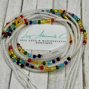 May include: White waist beads with colorful glass beads, including red, yellow, blue, and green. The beads are arranged in a layered pattern. The text "Luxe Adornments Co. Self Love & Manifestation Boutique" is visible on a white card.