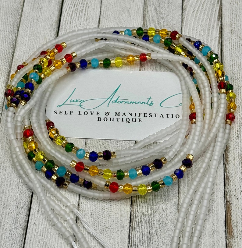 May include: White waist beads with colorful glass beads, including red, yellow, blue, and green. The beads are arranged in a layered pattern. The text "Luxe Adornments Co. Self Love & Manifestation Boutique" is visible on a white card.
