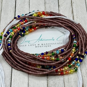 May include: A close-up of a brown beaded waist beads with colorful glass beads woven in. The beads are arranged in a rainbow pattern. The beads are on a white surface with the text "Luxe Adornments Self Love & Manifestation Boutique".