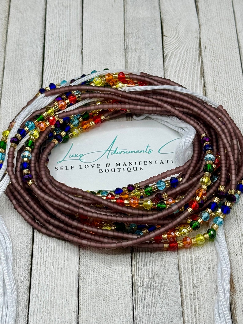 May include: A close-up of a brown beaded waist beads with colorful glass beads woven in. The beads are arranged in a rainbow pattern. The beads are on a white surface with the text "Luxe Adornments Self Love & Manifestation Boutique".