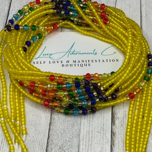 May include: Yellow beaded waist beads with rainbow-colored accents. The beads are strung together and tied with white string. The image includes a card with the text "Luxe Adornments Co. Self Love & Manifestation Boutique."