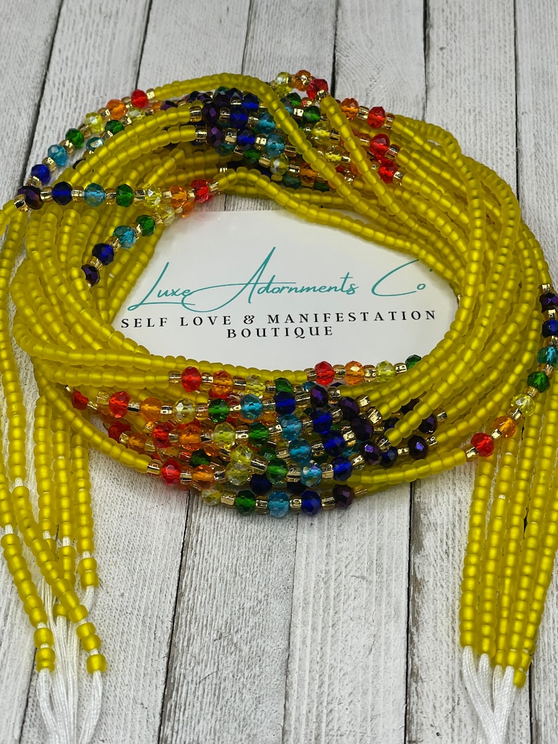 May include: Yellow beaded waist beads with rainbow-colored accents. The beads are strung together and tied with white string. The image includes a card with the text "Luxe Adornments Co. Self Love & Manifestation Boutique."