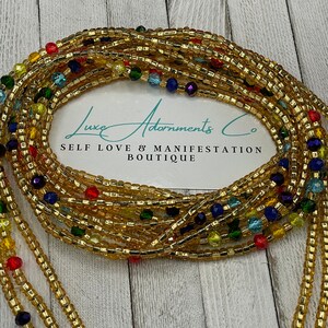 Puede incluir: Cuentas de cintura doradas con cuentas de acento coloridas. Las cuentas est&aacute;n dispuestas en m&uacute;ltiples hebras y est&aacute;n drapeadas sobre una tarjeta blanca con el texto "Luxe Adornments Co" y "Self Love & Manifestation Boutique".