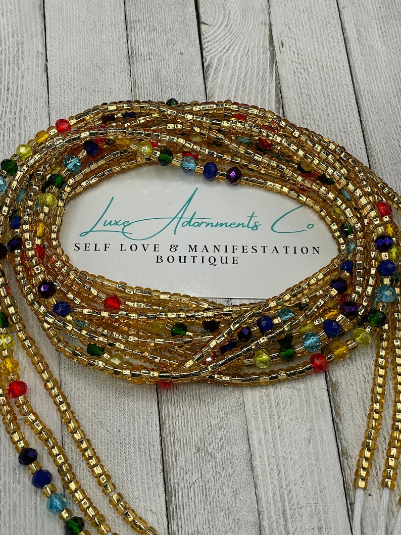 Puede incluir: Cuentas de cintura doradas con cuentas de acento coloridas. Las cuentas est&aacute;n dispuestas en m&uacute;ltiples hebras y est&aacute;n drapeadas sobre una tarjeta blanca con el texto "Luxe Adornments Co" y "Self Love & Manifestation Boutique".