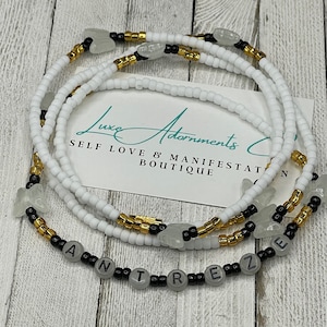 May include: A white and black beaded waist beads with gold accents. The beads spell out the word "ANTIFREEZE".