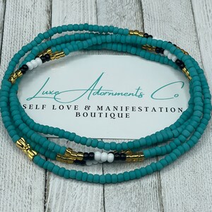 May include: A turquoise, black, white, and gold beaded waist bead necklace. The beads are strung on a thin cord and are arranged in a repeating pattern. The necklace is on a wooden surface. The text "Luxe Adornments Co. Self Love & Manifestation Boutique" is visible on a white card behind the necklace.