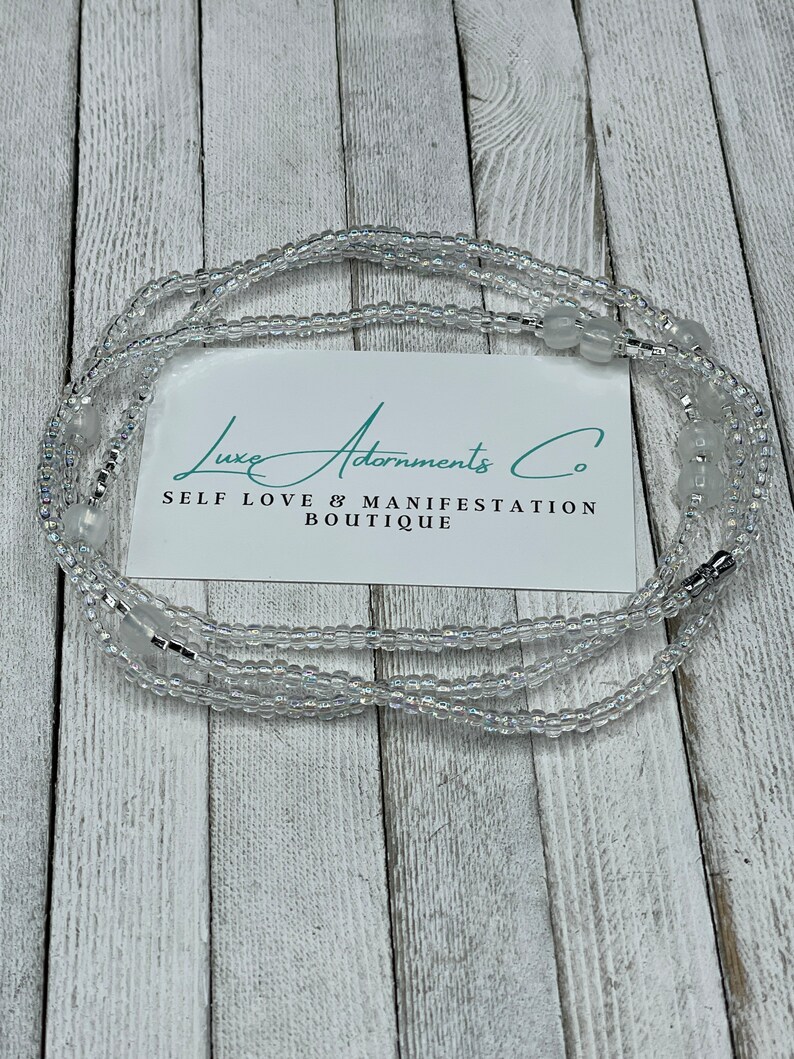 May include: A white beaded waist chain with silver accents and a card that reads "Luxe Adornments Co. Self Love & Manifestation Boutique."