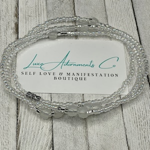 May include: A clear beaded waist chain with silver accents and white beads. The chain is on a wooden surface with a white card that reads "Luxe Adornments Co. Self Love & Manifestation Boutique".