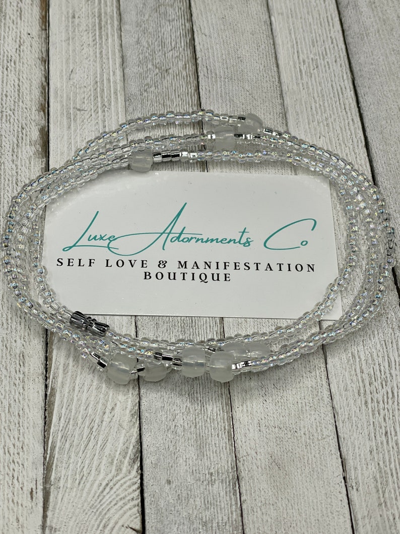 May include: A clear beaded waist chain with silver accents and white beads. The chain is on a wooden surface with a white card that reads "Luxe Adornments Co. Self Love & Manifestation Boutique".