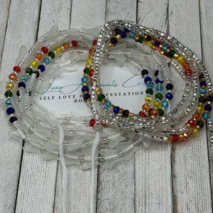 May include: Two strands of waist beads with colorful beads and clear beads. The beads are arranged in a rainbow pattern. The beads are on a white surface with the text "Luxe Adornments Co. SELF LOVE & BODY MANIFESTATION".