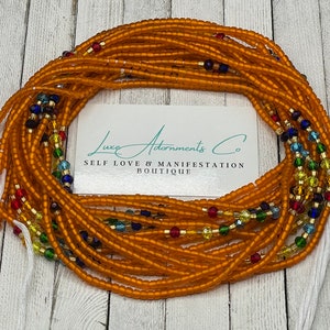 May include: A waist bead necklace made with amber-colored glass beads and colorful glass accents. The beads are strung on a white cord. The necklace is displayed on a wooden surface with a business card that reads "Luxe Adornments Co. Self Love & Manifestation Boutique."