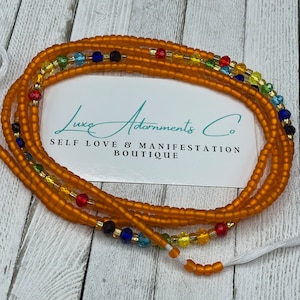 K&ouml;nnte beinhalten: Eine mehrstr&auml;ngige Perlenkette mit brauner Basis und bunten Akzenten. Die Kette besteht aus kleinen, runden Perlen und hat einen Quastenanh&auml;nger am Ende. Der Text "Luxe Adornments Co. Self Love & Manifestation Boutique" ist auf einer wei&szlig;en Karte hinter der Kette sichtbar.