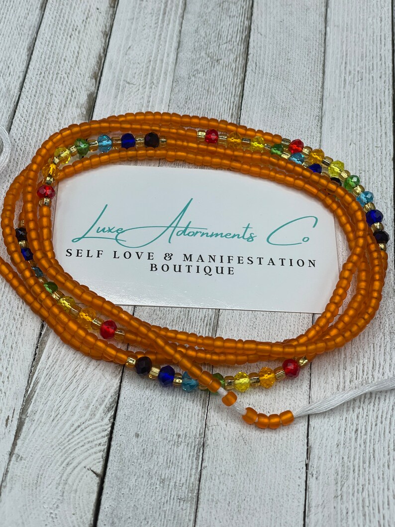 K&ouml;nnte beinhalten: Eine mehrstr&auml;ngige Perlenkette mit brauner Basis und bunten Akzenten. Die Kette besteht aus kleinen, runden Perlen und hat einen Quastenanh&auml;nger am Ende. Der Text "Luxe Adornments Co. Self Love & Manifestation Boutique" ist auf einer wei&szlig;en Karte hinter der Kette sichtbar.