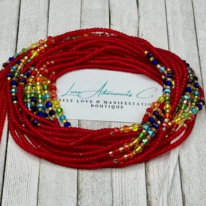 May include: Red waist beads with rainbow-colored crystal accents and gold-colored beads. The beads are arranged in multiple strands. The image includes a white card with the text "Luxe Adornments Co. SELF LOVE & MANIFESTATION BOUTIQUE."