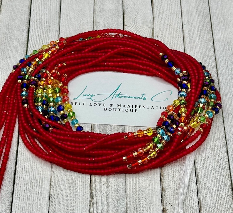 May include: Red waist beads with rainbow-colored crystal accents and gold-colored beads. The beads are arranged in multiple strands. The image includes a white card with the text "Luxe Adornments Co. SELF LOVE & MANIFESTATION BOUTIQUE."