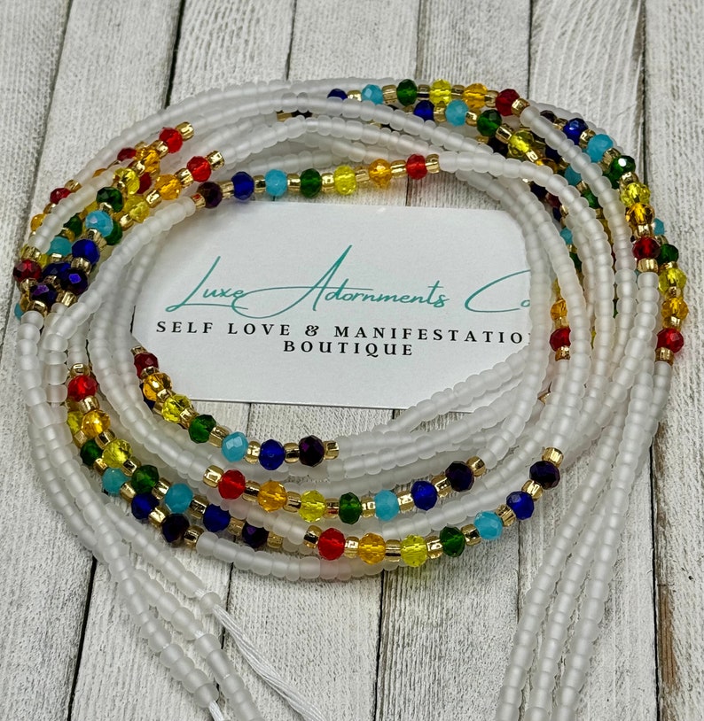 May include: White beaded waist beads with colorful crystal accents in red, yellow, green, blue, and purple. The beads are strung together, with a tag that reads "Luxe Adornments Co. SELF LOVE & MANIFESTATION BOUTIQUE."