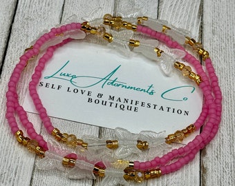 May include: Pink and gold beaded bracelet with clear butterfly charms. The bracelet is layered with three strands of beads and has the text "Luxe Adornments Co. Self Love & Manifestation Boutique" on a white card.