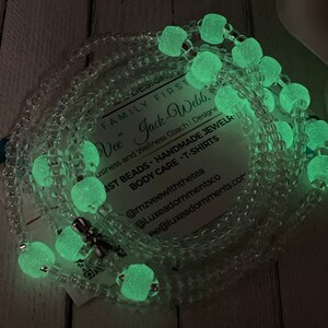 May include: A close-up of a glow-in-the-dark beaded necklace with a silver charm. The necklace is made of clear beads with green glow-in-the-dark beads interspersed throughout. The necklace is lying on a wooden surface. The text on the card reads: "FAMILY FIRST Vee" Jack-Webb, Business and Wellness Coach | Designe AST BEADS HANDMADE JEWELR BODY CARE -T-SHIRTS @mzveewiththetea @Luxeadornmentsco e@luxeadornments.com"