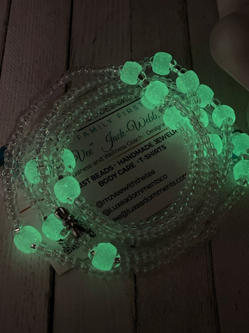 May include: A close-up of a glow-in-the-dark beaded necklace with a silver charm. The necklace is made of clear beads with green glow-in-the-dark beads interspersed throughout. The necklace is lying on a wooden surface. The text on the card reads: "FAMILY FIRST Vee" Jack-Webb, Business and Wellness Coach | Designe AST BEADS HANDMADE JEWELR BODY CARE -T-SHIRTS @mzveewiththetea @Luxeadornmentsco e@luxeadornments.com"