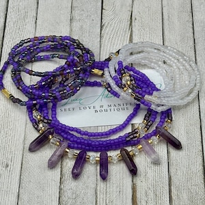 May include: An assortment of beaded waist beads in shades of purple, gold, and white. Some strands feature amethyst crystal pendants. The image includes a card with the text "SELF LOVE & MANIFEST BOUTIQUE".