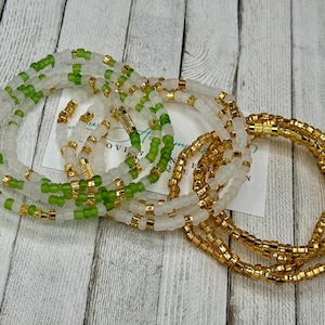 May include: A set of three beaded bracelets in white, green, and gold. The bracelets are made of small beads and are layered on top of each other.